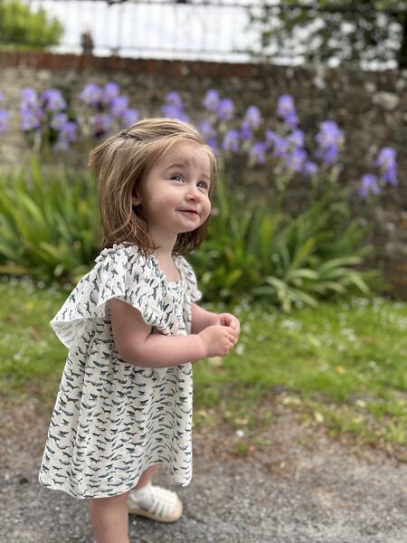 toddler standing in sunny garden wearing white dress in cotton jersey with frill over shoulders in whale print