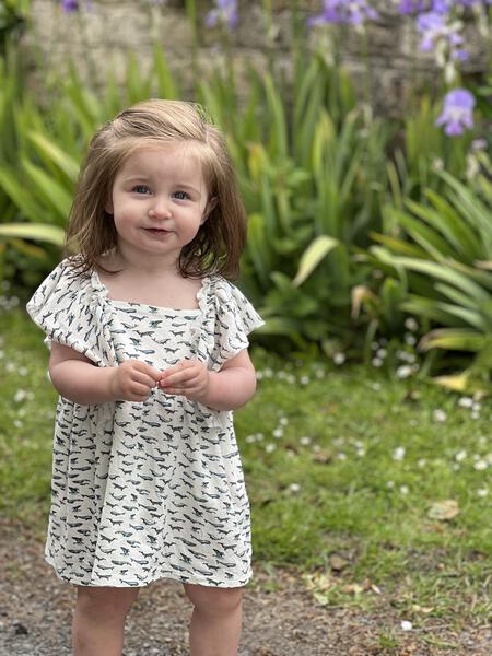 toddler standing in sunny garden wearing white dress in cotton jersey with frill over shoulders in whale print