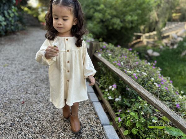 toddler standing in sunny garden wearing cream long sleeved jersey rib dress. buttons down the front