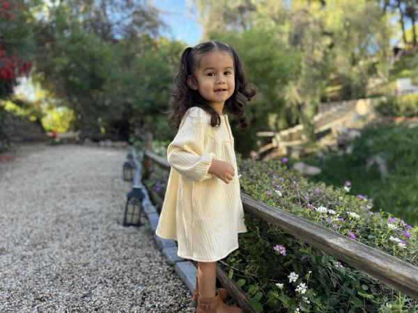 happy toddler standing in sunny garden wearing cream long sleeved jersey rib dress. buttons down the front