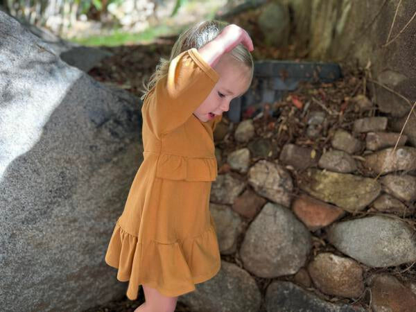 happy toddler in autumn sunshine wearing gold long sleeved dress with frill at waist and hem