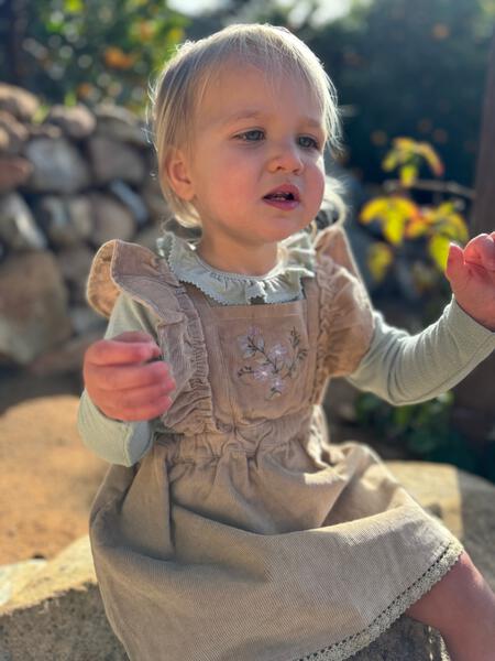 toddler in sunshine wearing green long sleeve patterned fabric tee with frill neck under overall pinny dress 