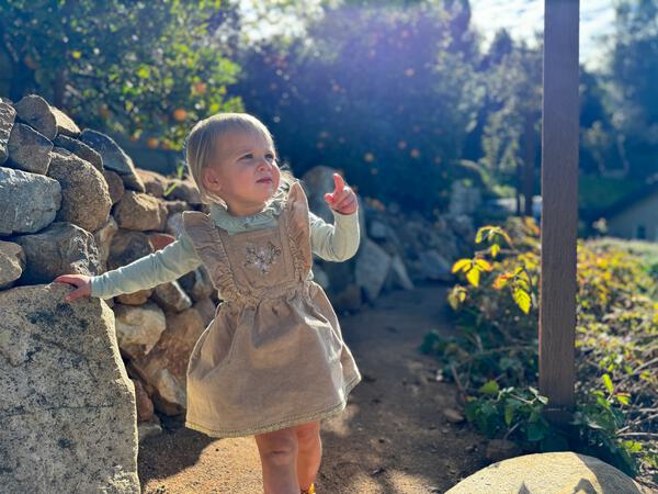 toddler in sunshine wearing green long sleeve patterned fabric tee with frill neck under overall pinny dress 