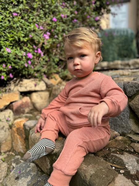 toddler sat in garden wearing rust sweatshirt with hands through patch pocket on the front side snap opening on neck and matching sweat pants