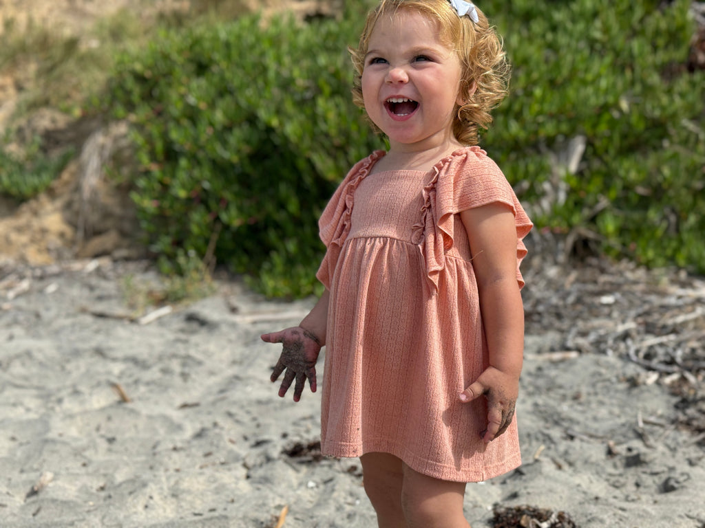 happy toddler laughing on beach on sunny day wearing coral dress in pointelle jersey with frill over shoulders