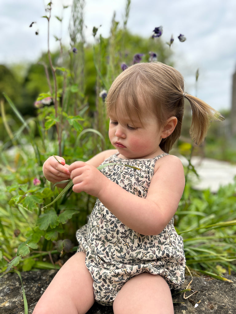 happy toddler sat on stone wall on sunny day wearing cream bubble with shoe string straps in flower and bird print