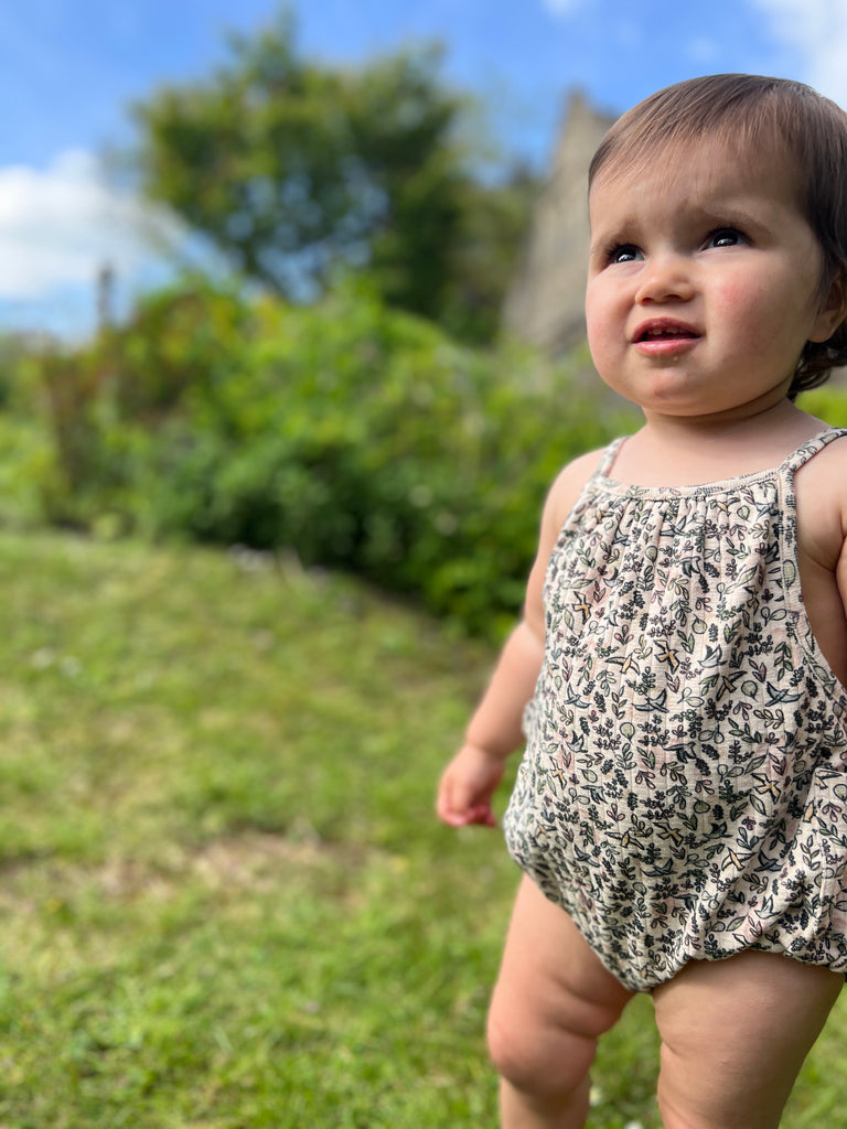 toddler standing on grass on sunny day wearing cream bubble with shoe string straps in flower and bird print 