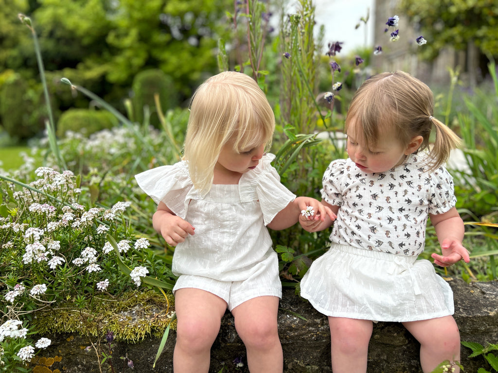 toddler sat on wall in sunny garden wearing short sleeved white ribbed jersey onesie with seahorse print and white skort friend wearing white romper