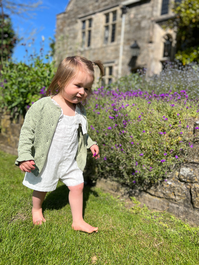 happy toddler standing in front of flowers wearing sage green lace knit cardigan frill cuffs on sleeve and white shortie jump suit