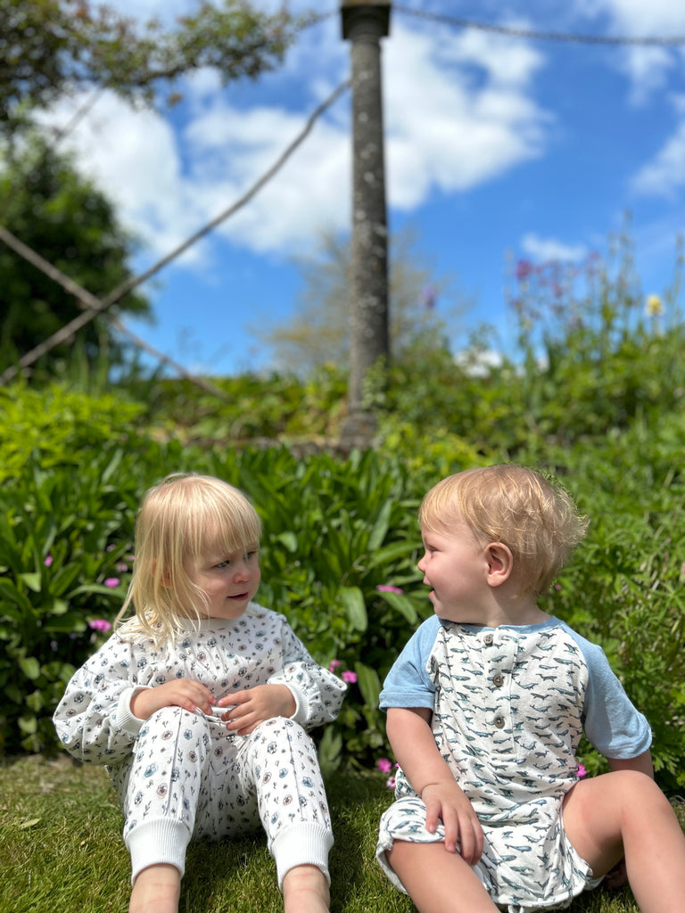 toddler sat on grass on sunny day with a friend wearing white flower print joggers with elastic waistband and drawcord side pockets and front seam detail and matching top