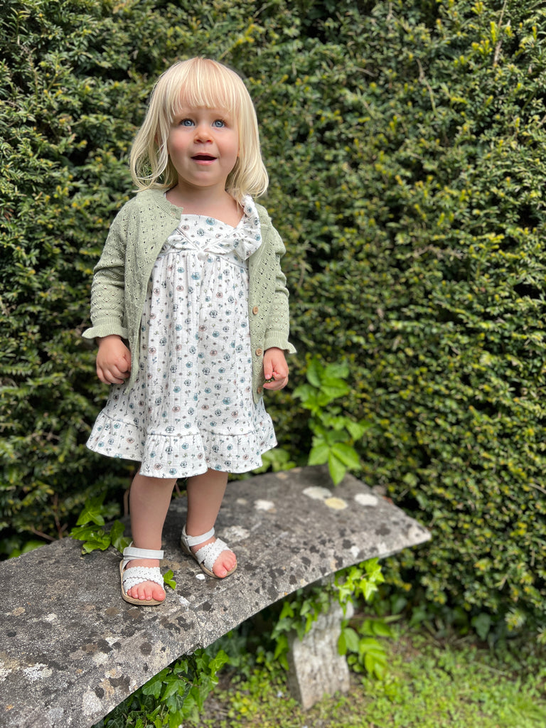 girl standing on old stone bench on sunny day wearing white dress with flower print frill shoulders hem frill and green knit cardigan