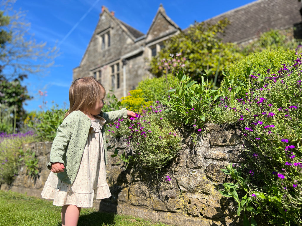 happy toddler standing in front of flowers wearing sage green lace knit cardigan frill cuffs on sleeve and cream dot dress