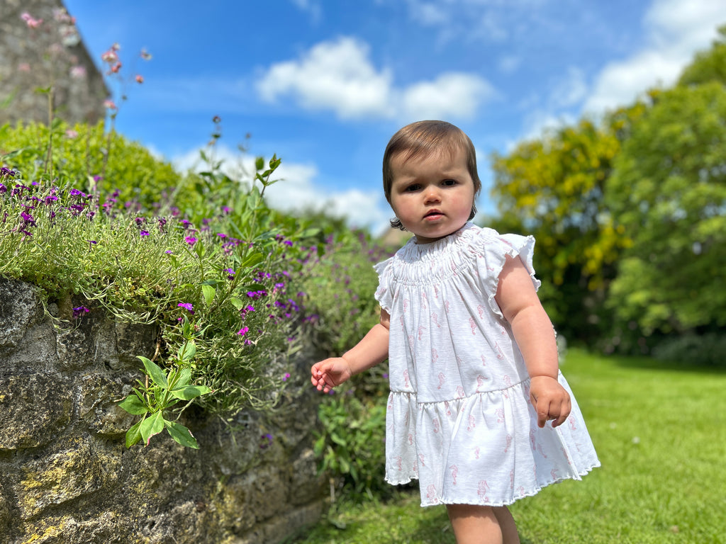 toddler standing in sunny garden in front of summer flowers wearing white dress with pink flower print smocked neck line short sleeves and large frill tier