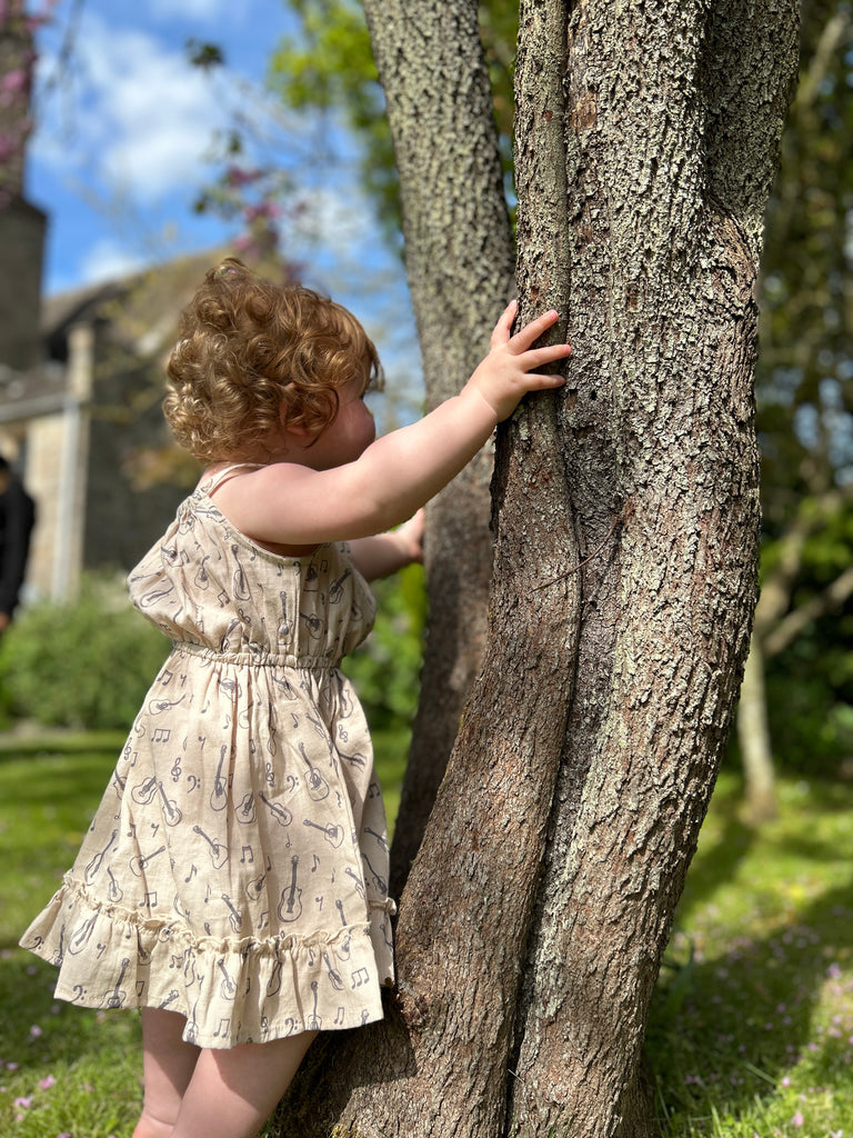 girl stood by tree in summer garden  on sunny day wearing cream dress with guitar print elastic waist shoestring shoulder straps hem frill