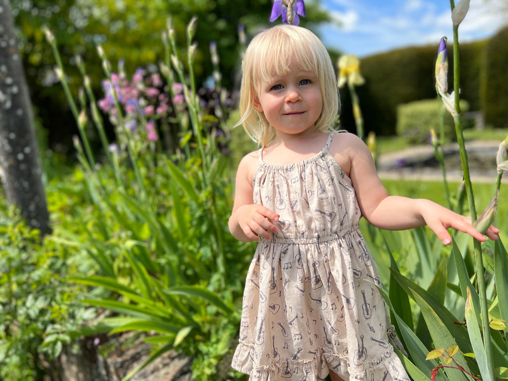 girl stood in summer garden flowers on sunny day wearing cream dress with guitar print elastic waist shoestring shoulder straps hem frill