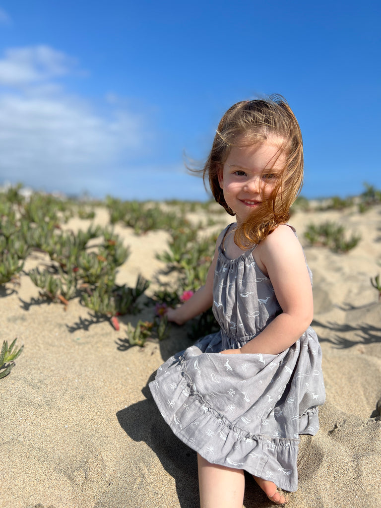 girl sat on beach on sunny day wearing grey dress with whale print elastic waist shoestring shoulder straps hem frill 