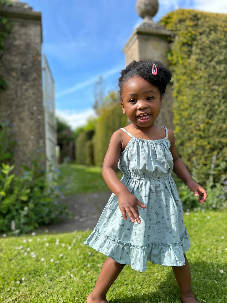 girl in sunny garden wearing blue dress with island and palm tree print elastic waist shoestring shoulder straps hem frill