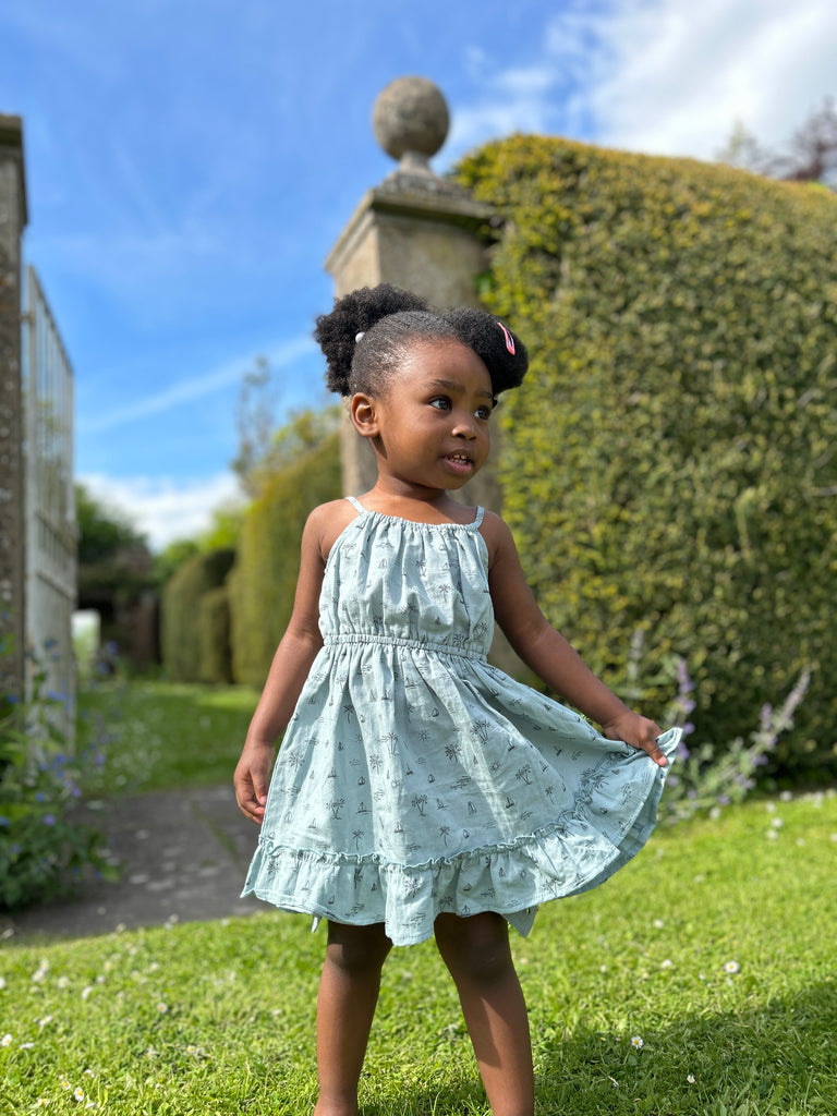 girl in sunny garden wearing blue dress with island and palm tree print elastic waist shoestring shoulder straps hem frill