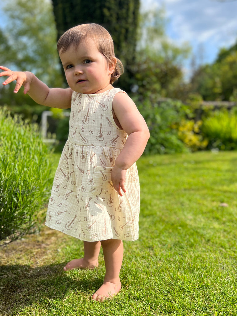 toddler in sunny garden wearing cream gauze sleeveless dress with all over brown guitar print 2 front patch pockets