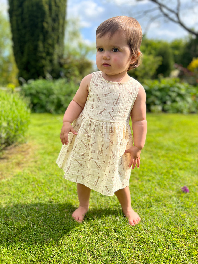 toddler in sunny garden wearing cream gauze sleeveless dress with all over brown guitar print 2 front patch pockets