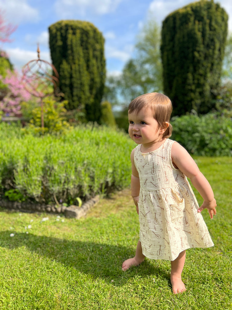 toddler in sunny garden wearing cream gauze sleeveless dress with all over brown guitar print 2 front patch pockets