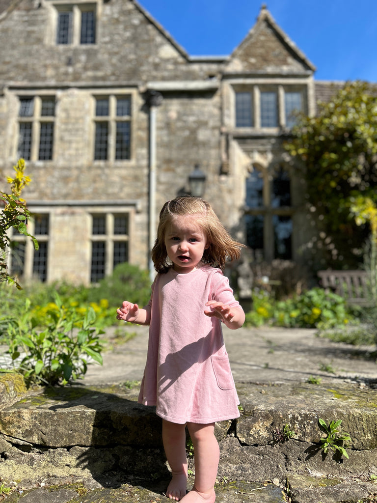 toddler standing on old stone pathway in front of old manor house wearing short sleeve A-line soft pink dress with side patch pockets and round neck