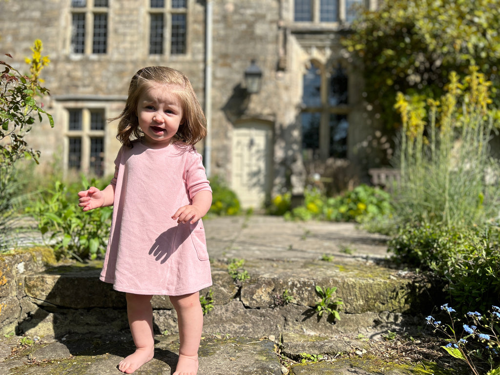 toddler standing on old stone pathway in front of old manor house wearing short sleeve A-line soft pink dress with side patch pockets and round neck