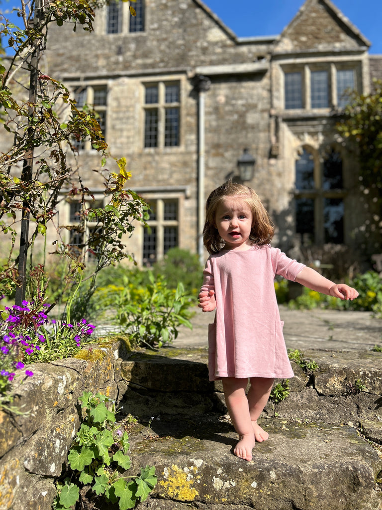 toddler standing on old stone pathway in front of old manor house wearing short sleeve A-line soft pink dress with side patch pockets and round neck