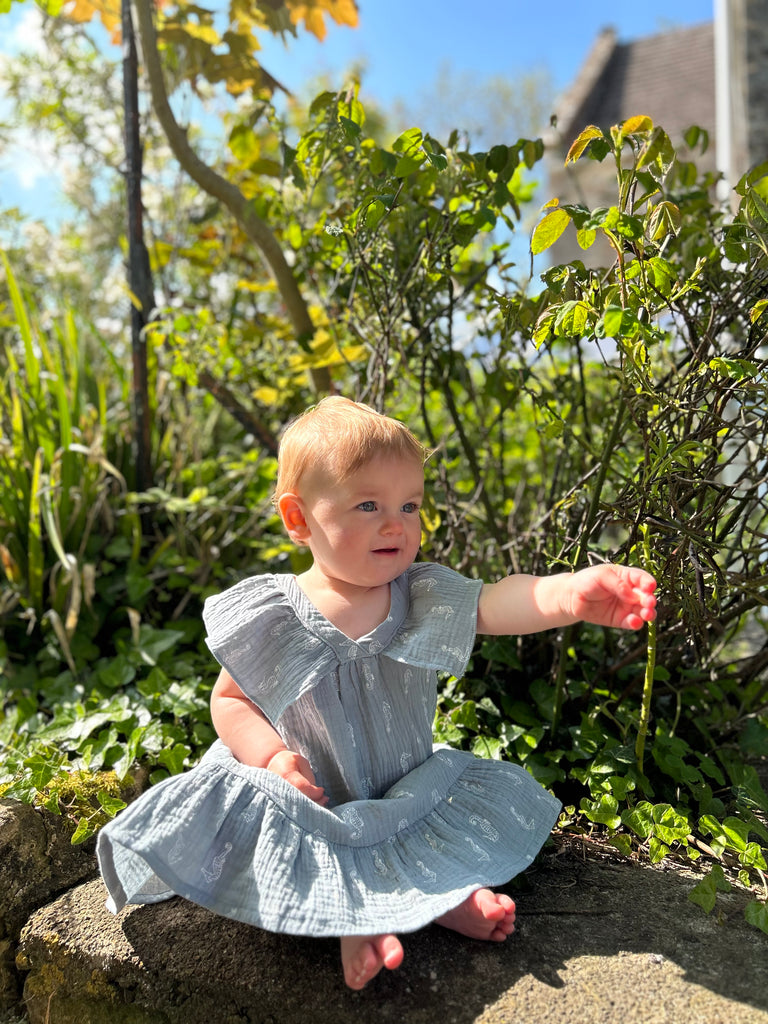 happy baby sat on a rock in sunny garden wearing delicate blue gauze summer dress with white seahorse print. Large over the shoulder frills and v neck front and back