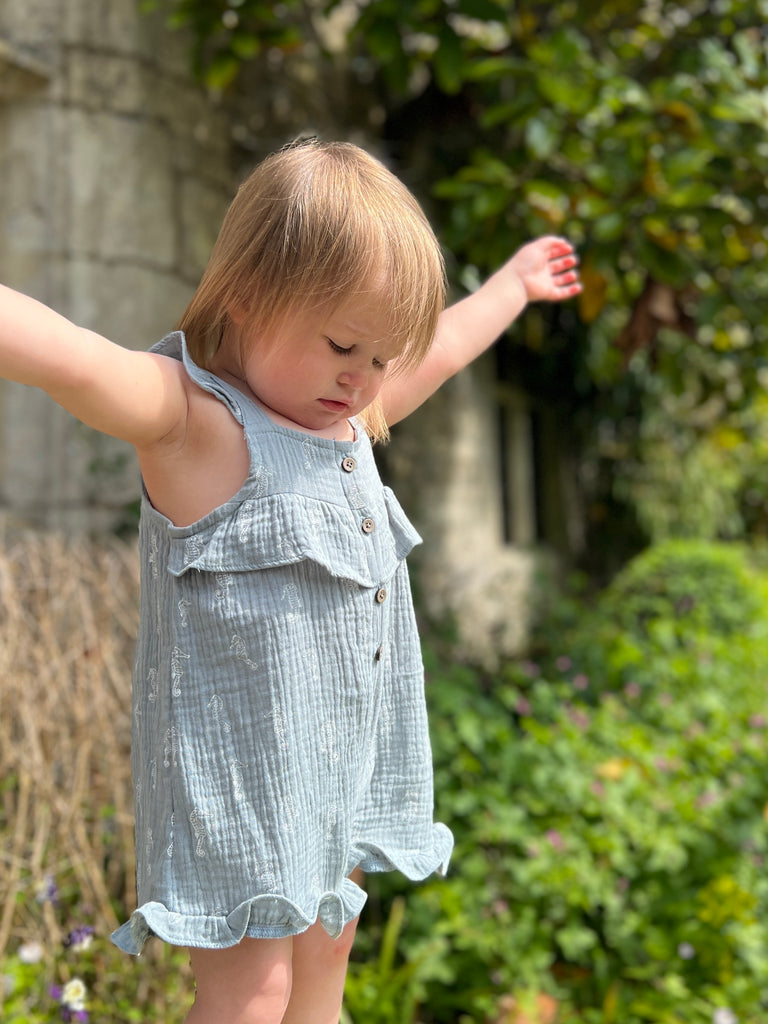 toddler standing in front of old stone window of manor house with arms in the air wearing gentle blue gauze shortie overalls with white all over seahorse print. buttons on front and frill detail round front and legs