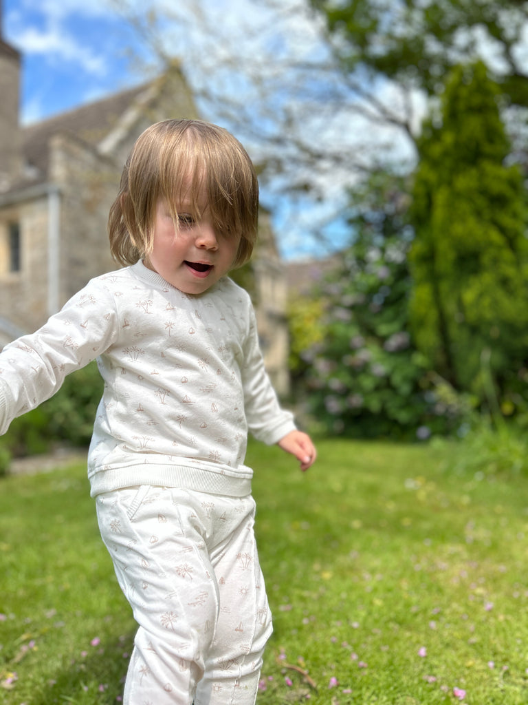 boy in garden on sunny day wearing white sweatshirt with all over cream island and boat print poppers at neck  and matching pants