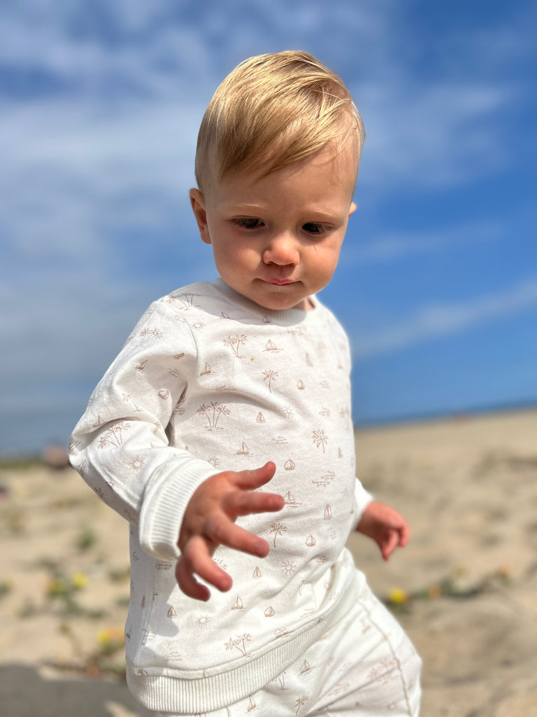 toddler on beach on sunny day with big blue sky wearing white sweatshirt with all over cream island and boat print poppers at neck and matching pants