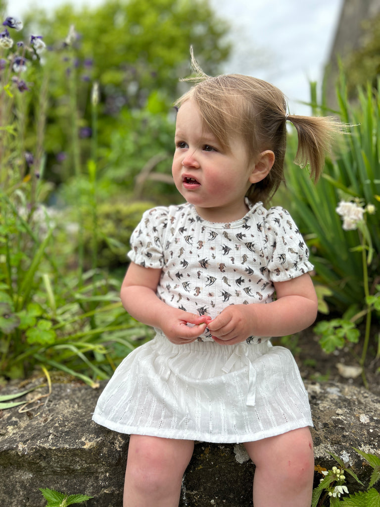 toddler sat on wall in sunny garden wearing short sleeved white ribbed jersey onesie with seahorse print and white skort