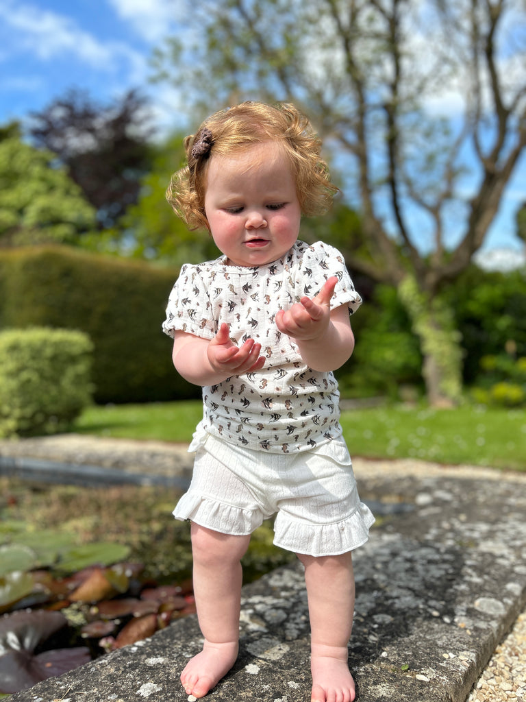 toddler in sunny garden wearing white gauze shorts with 2 side pockets and frill around legs with white tee