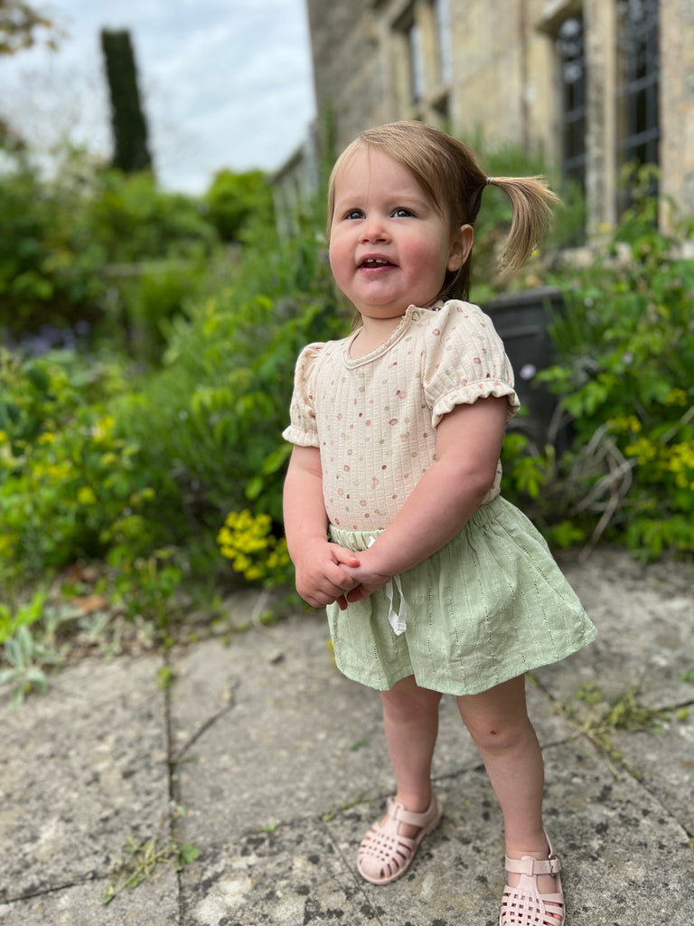 toddler standing in sunny garden in front of old stone house wearing short sleeved cream ribbed jersey onesie with pastel dot print and green skirt