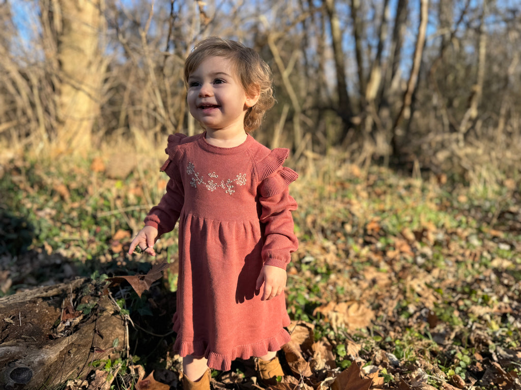 toddler in autumnal woods wearing rust long sleeved cotton knit dress with flower embroidered panel, frill sleeves and hem