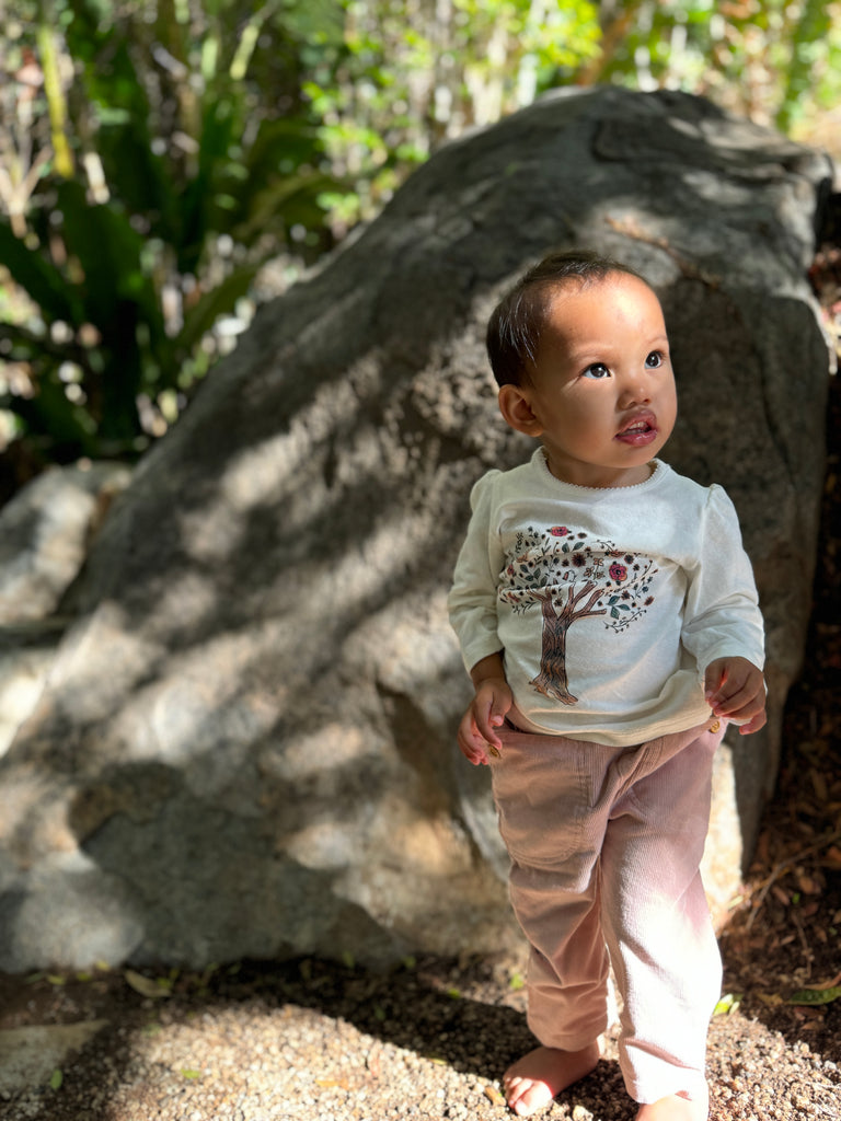 happy toddler in sunny garden wearing pink cord pants with patch pockets and button detail and tree print tee