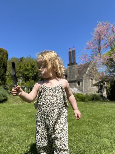 happy toddler in sunny garden wearing  jersey long legged romper with shoe string straps and frills Small flowers and birds print on cream background