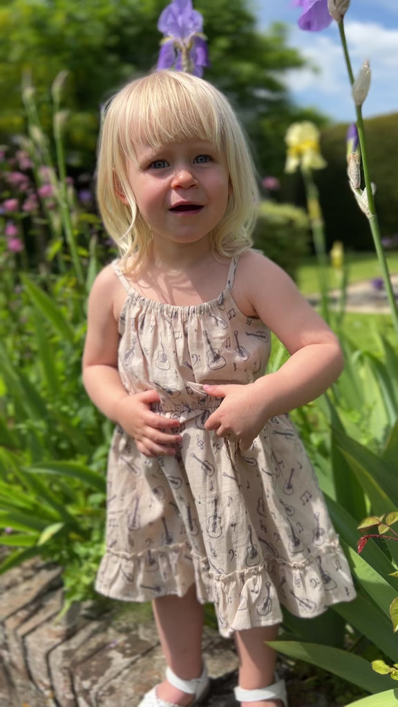 girl stood in summer garden flowers on sunny day wearing cream dress with guitar print elastic waist shoestring shoulder straps hem frill