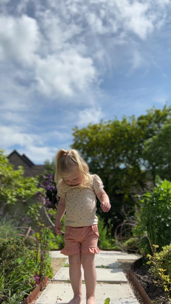 toddler standing on garden path in front of old stone house wearing tee shirt in cream ribbed cotton with pastel dotty print and coral shorts