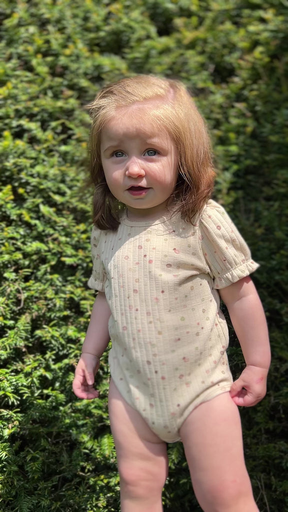 toddler standing in sunny garden in front of green hedge wearing short sleeved cream ribbed jersey onesie with pastel dot print