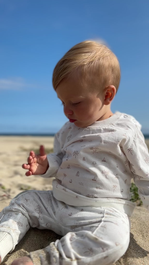 toddler on beach on sunny day with big blue sky wearing white sweatshirt with all over cream island and boat print poppers at neck and matching pants