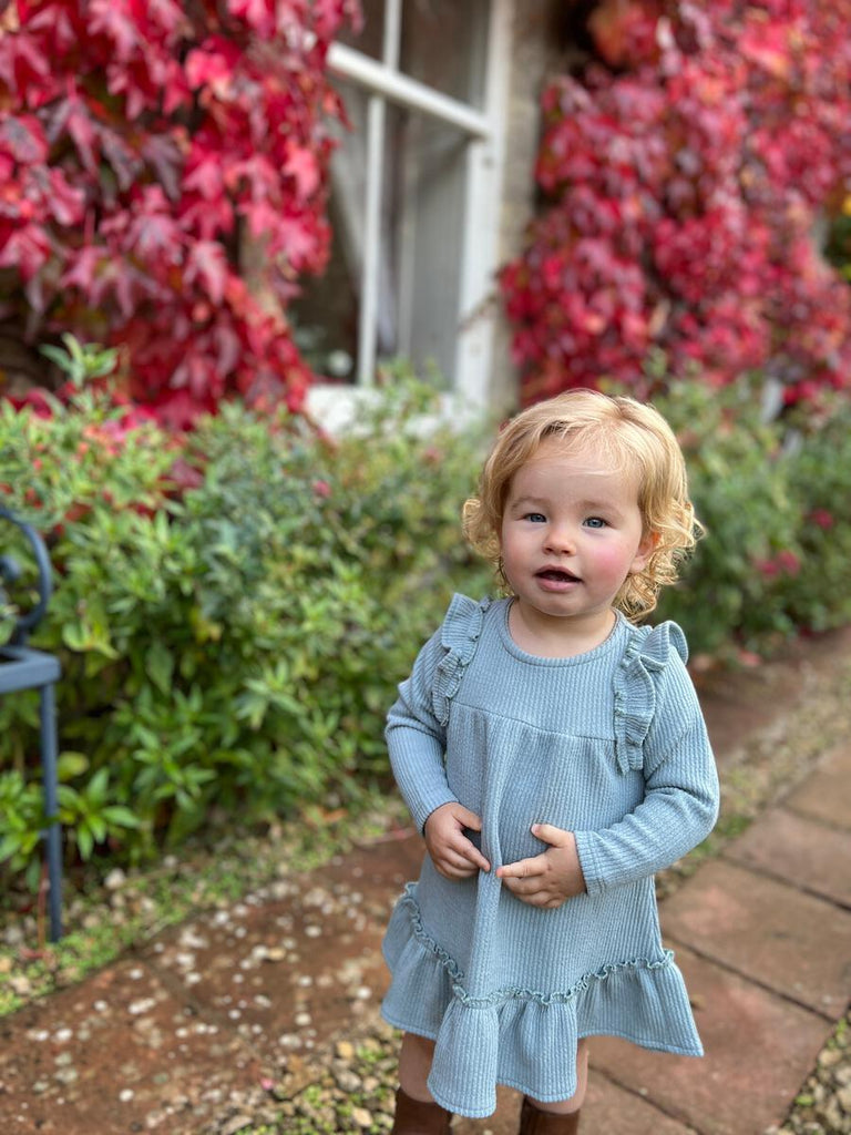 toddler girl standing on path wearing blue ribbed long sleeve dress with ruffles over the shoulders and tiered frill on bottom of dress