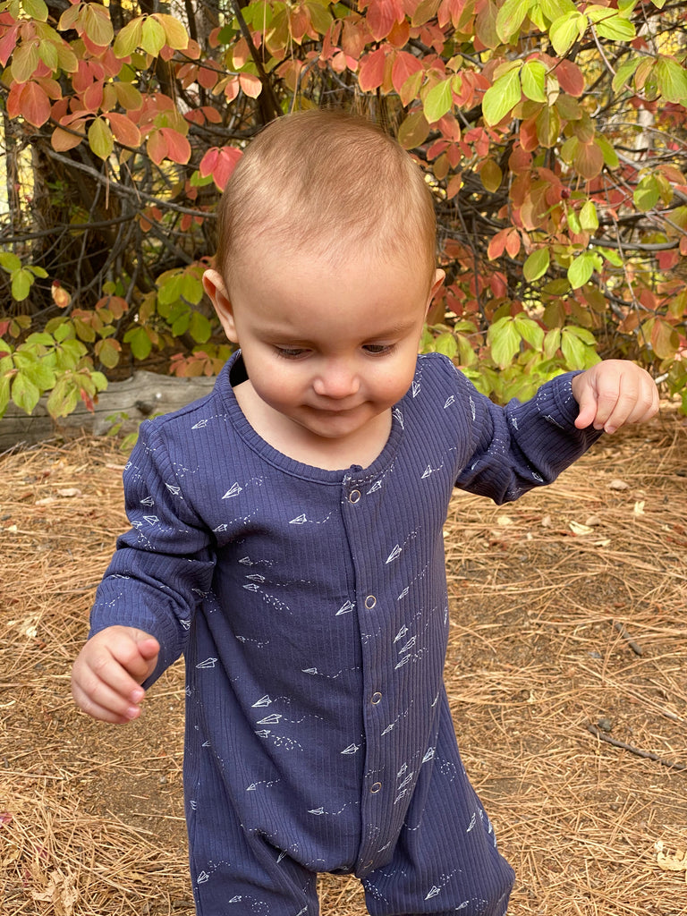 baby stood in front of autumn bush wearing navy blue ribbed romper with rocket print. poppers down front