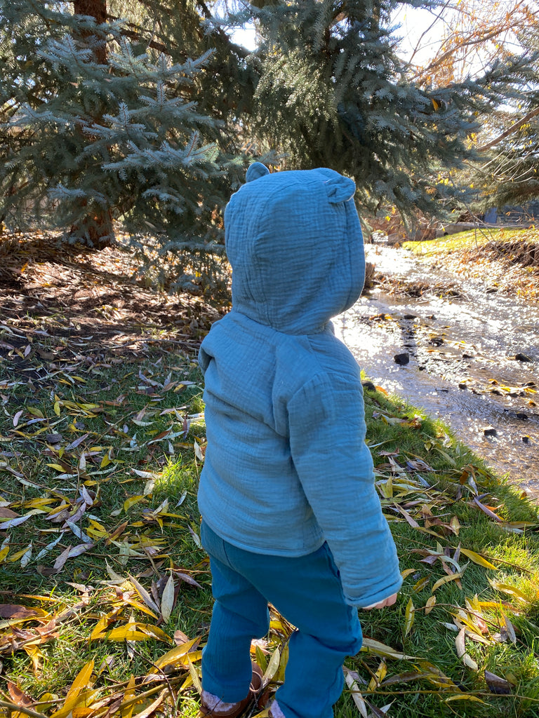 toddler standing in autumnal wood near stream wearing padded jacket zip front with hood teal green jersey with leaf print lining bear ears on hood