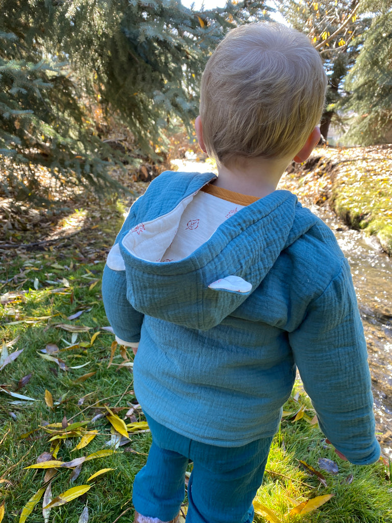 toddler standing in autumnal wood near stream wearing padded jacket zip front with hood teal green jersey with leaf print lining bear ears on hood